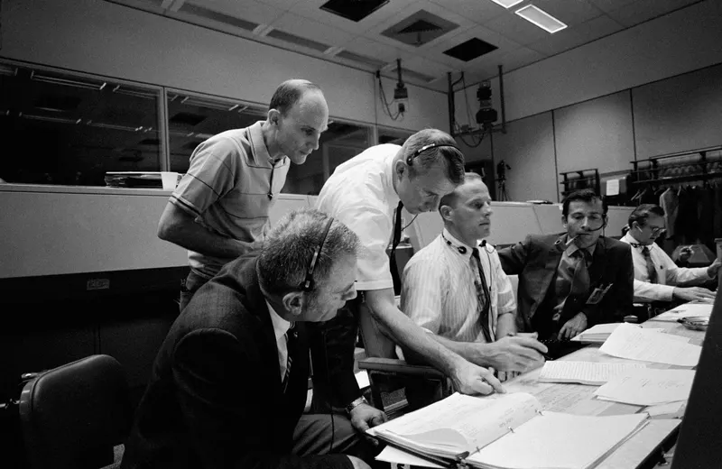 Black and white photo of flight controllers huddled over a console in Mission Control, Houston, during Apollo 13.